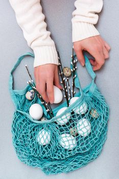 Happy Easter. Female hands put chicken and quail eggs and pussy willow branches in a blue string bag on a gray background