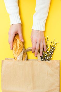 Food delivery concept. Female hands hold a paper bag with bread and a bouquet of pussy willow on a yellow background. Vertical view