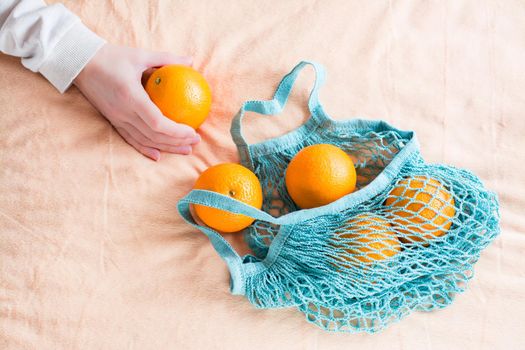 Woman's hand folds fresh oranges in a mesh bag on a fabric background. Zero waste