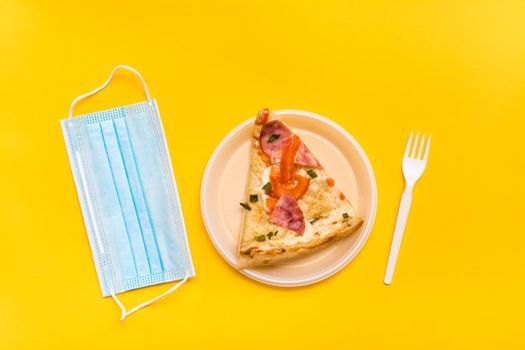 Takeaway and delivery. A slice of pizza in a disposable plastic plate, a protective mask and a fork on a yellow background