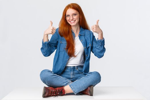 Excellent, satisfaction and people concept. Delighted supportive cute redhead girlfriend sitting on floor, with legs crossed in relaxed pose, smiling approval, showing thumbs-up cheering.