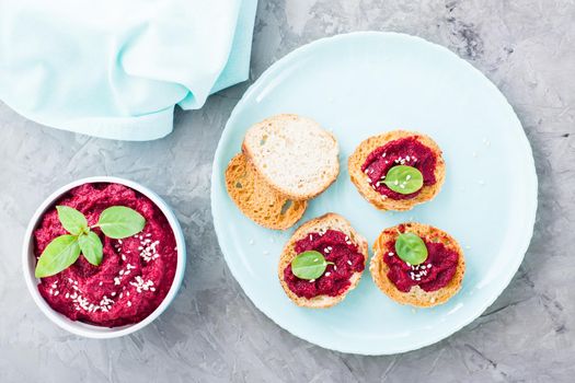 Homemade bruschetta with beetroot hummus on small baked baguette toast on a plate and a bowl of hummus on the table. Top view