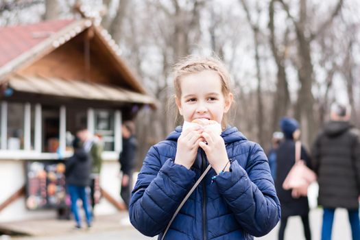 A cute girl takes a bite of a pie bought in a food truck in a city park in early spring. Takeaway food