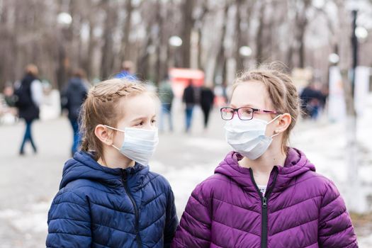 Two girls in protective face masks in the park on the background of a crowd of people. Walk the new normal and social distance in a pandemic