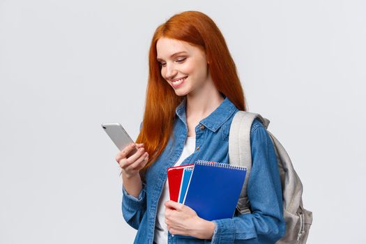 Modern lifestyle, technology and education concept. Cute, lovely redhead female student heading to campus after classes in college, holding backpack and notebooks, chatting on smartphone.