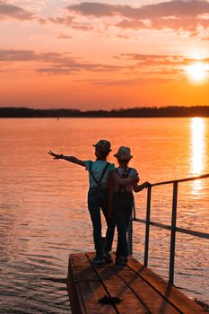 Two girls sisters stand on a wooden bridge near the river bank at sunset and look into the distance. Local tourism