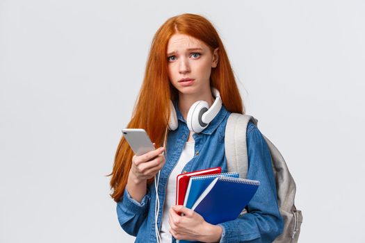 Uneasy, upset and gloomy cute redhead teenage girl, student in university frowning look distressed and unhappy camera, holding backpack education notebooks and smartphone.