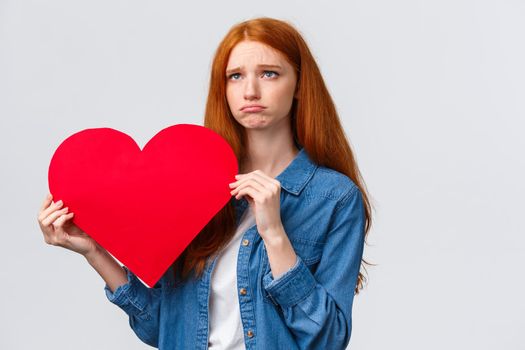 Unhapy brokenhearted cute redhead girl, sulking and looking up uneasy, distressed holding big red valentines day heart, was rejected, showed her feelings standing white background.