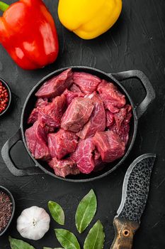 Fresh beef chunks served on table with ingredients ready for cooking set with sweet bell pepper, in cast iron frying pan, on black stone background, top view flat lay