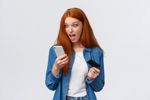 Excited and shocked young redhead girl hurry up buy dress with awesome discount, staring impressed and surprised smartphone display, holding mobile phone and credit card, white background.