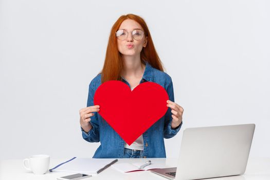 Lovely, romantic cute redhead girl folding lips in mwah, send air kiss and holding big red valentines card, showing her love and passion, congratulate girlfriend with anniversary, stand near laptop.