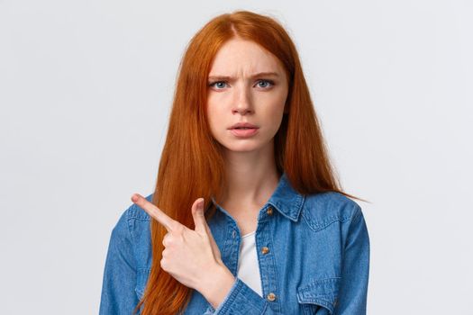 Close-up portrait serious-looking skeptical and displeased angry bothered redhead girlfriend waiting explanation, demand answers as seing something unplesant, pointing finger left.