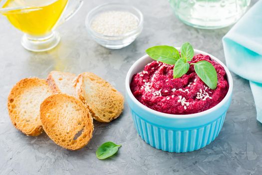 Homemade baked beet hummus in a bowl with sesame seeds and basil and baked small toast on the table