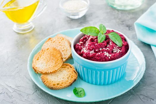 Homemade baked beetroot hummus in a bowl with sesame seeds and basil and baked small toast on a plate on the table