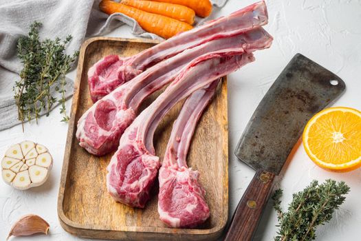 Raw lamb chops or mutton cuts set, with ingredients carrot orange, herbs, and old butcher cleaver knife, on white stone background