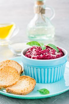 Homemade baked beetroot hummus in a bowl with sesame seeds and basil and baked small toast on a plate on the table. Vertical view