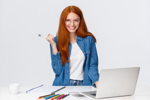 Sassy and confident good-looking charismatic redhead female near laptop and table with drawing supplies, smiling cheeky and mystic camera, standing white background have excellent plan.