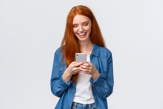 Chatting, communication and mobile concept. Cheerful redhead smiling girl texting friend, laughing as watching funny vlog, looking smartphone display happy, standing white background.