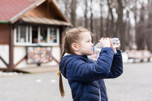 Cute little girl drinks water from a bottle on the background of a food truck in a city park. Takeaway food