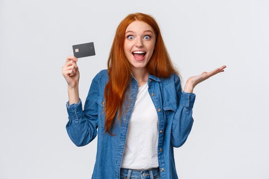 Waist-up portrait surpirsed and cheerful redhead girl talking about bank service, holding credit card smiling amused and excited, raise hand in wonder and thrill, standing white background.