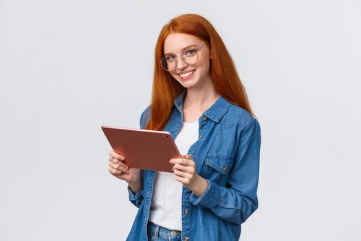 Professionalism, work and people concept. Young cheerful attractive female redhead in glasses, holding digital tablet and smiling at camera with happy confident expression over white background.