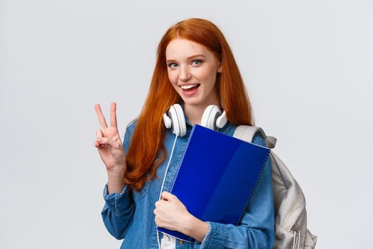 Lovely friendly college student, redhead girl with papers, documents for school, holding backpack and wear headphones over neck, make peace gesture informal greeting classmate, white background.