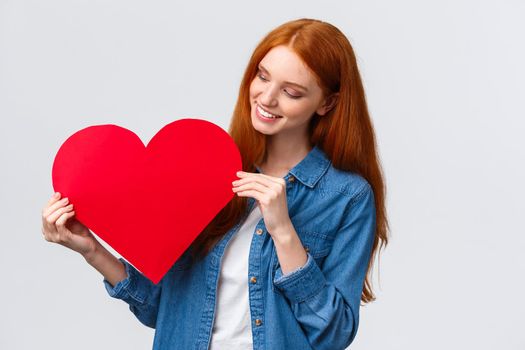 Valentines day, sympathy and relationship concept. Lovely creative cute redhead girl made red big heart for gift, holding cardboard and smiling tender, confess love and passion, white background.