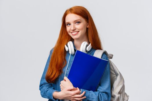 Charismatic lovely teenage college girl, redhead female student with notebooks and papers, prepare project for todays class, wear headphones over neck and backpack, white background.