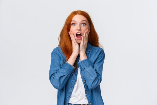 Oh my gosh, amazing. Surprised speechless redhead woman standing starled and shocked, touch face, drop jaw and stare camera fascinated, got job, won prize, standing white background.