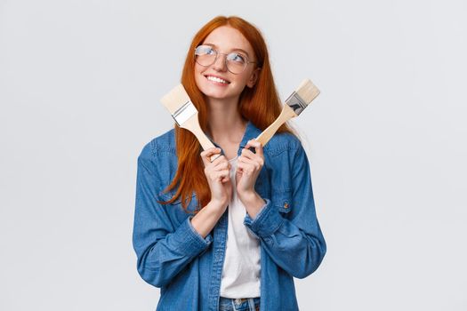 Creativity, imagination and people concept. Good-looking redhead dreamy girl designer, planning to renovate, paint walls in appartment, looking thoughtful and holding paintbrush, white background.