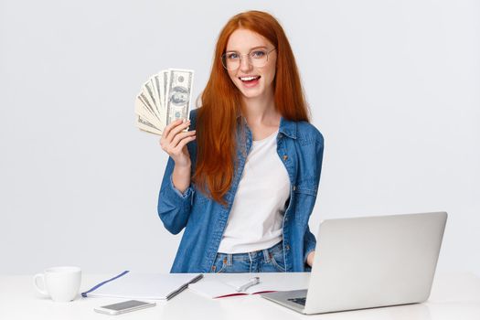 Girl likes her new work, got promoted and receive first paycheck. Attractive sassy redhead woman in glasses, standing near working table, laptop, holding money, big cash.