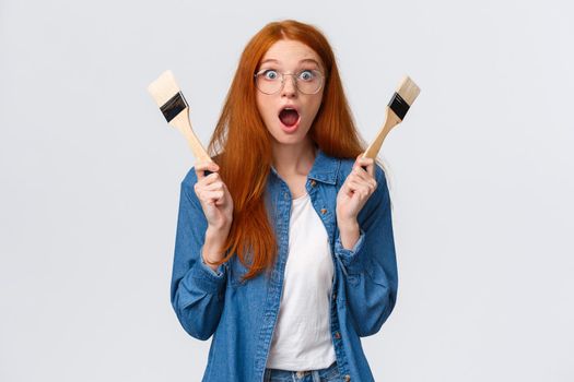 Amused and excited, wondered redhead woman creating new art, painting, look stunned and thrilled, say wow drop jaw and pop eyes camera, holding two paintbrushes, white background.