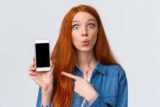 Curious and amused lovely redhead woman with long red hair, folding lips intrigued and excited, discussing new app, photos of classmate with new car, pointing finger smartphone, gossiping.