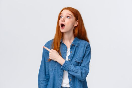 Waist-up portrait amazed, speechless pretty redhead teenage collage student, ginger long hair, drop jaw gasping and looking upper left corner wondered, pointing at fascinating product.