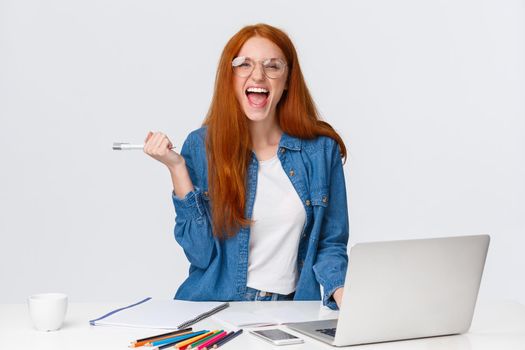 Excited and triumphing good-looking redhead female student passed challenge, found out final exam rating online using laptop, holding pen and screaming happily, standing white background.