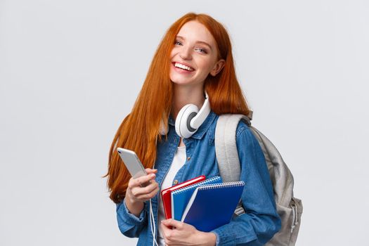 Cheerful, friendly and charismatic redhead woman laughing joyfully, texting friends, chatting with smartphone, enjoying college lifestyle, heading campus with backpack and notebooks.