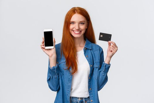 Waist-up portrait confident female with red long hair, blue eyes, holding credit card and showing smartphone display, easy banking service, payment for bills internet, smiling satisfied.