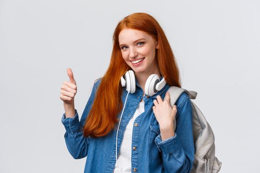 Girl found excellent part-time job after university classes. Attractive redhead woman with backpack, headphones over neck, showing thumb-up and smiling in approval, recommending.