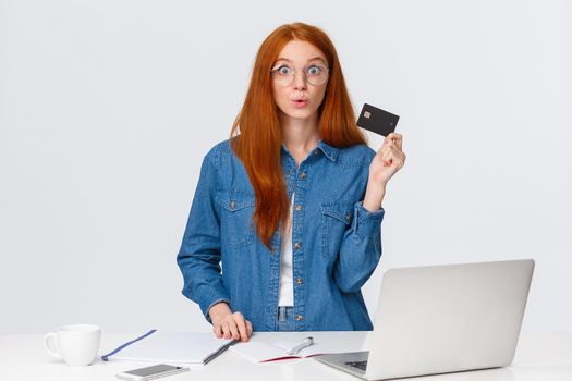 Excited and amused, curious good-looking redhead girl want try cashback online bank offer, holding credit card, folding lips in wow wondered, look camera impressed, stand near laptop.