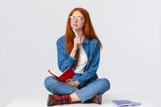 Concentrated, thoughtful and creative young female genius with red hair, in glasses, looking perplexed and thinking over solution to task, writing homework sitting on floor with notebook and pen.