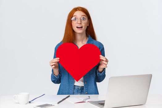 Amused and dreamy surprised redhead girl received big red valentines day heart from coworker, standing near working table with present, gasping astonished, white background.