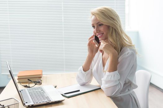 Business, technology and communication concept - blonde woman talking phone while watching laptop.