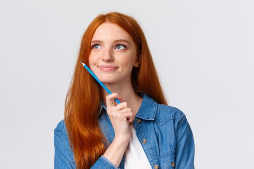 Close-up portrait inspired, dreamy and talented cute redhead woman thinking what draw, holding colored pencil and smiling pleased, imaging artwork, designing over white background.