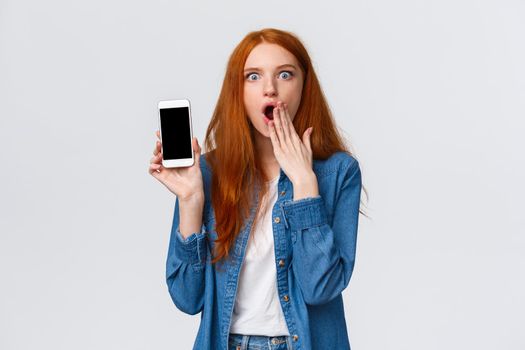 Impressed and astonished redhead woman seeing something unbelievable, spilling tea in internet, gasping cover opened mouth and showing samrtphone display, white background.