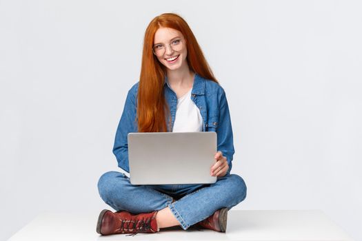 Cheerful sassy redhead female student, college girl in glasses, sitting on crossed legs with laptop, working freelance, prepare project awat from office, remote over white background.