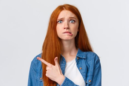 Anxious and worried concerned redhead caucasian girl having big problems, nervously biting lip and frowning scared, pointing afraid left, standing white background waiting be scolded.