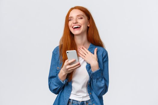 Waist-up portrait carefree and joyful, alluring redhead caucasian girl receive funny meme message, holding smartphone, laughing and close eyes dreamy, standing white background.