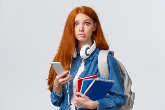 Indecisive, nervous cute redhead freshman girl, start new year college, looking worried camera, biting lip nervously, holding backpack, notebooks and smartphone, reading message.