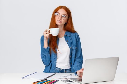 Dreamy, talented good-looking redhead girl studying design and art, looking dreamy and thoughtful up, creating new project, artwork, standing with cup coffee near laptop and pencils.