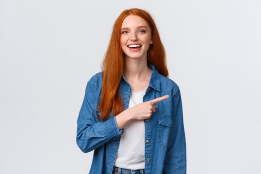 Carefree, friendly and happy outgoing redhead woman in denim shirt, pointing finger right and laughing at camera with beaming smile, standing white background, having discussion, communicate team.